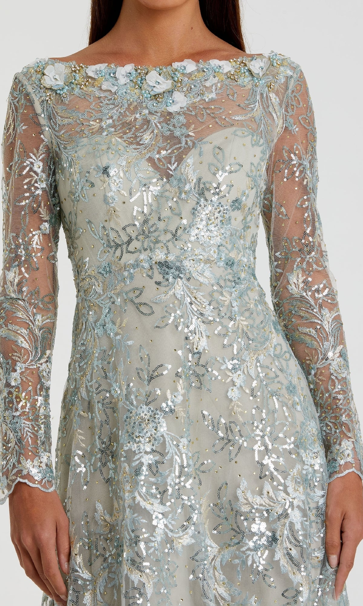 A woman wears the Mac Duggal 67498 long sleeve formal dress, featuring sheer fabric with silver and light blue floral embroidery and sequin details.